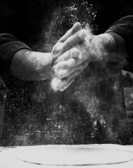 Black and white photo of a chef clapping flour in a kitchen, creating a dramatic effect.