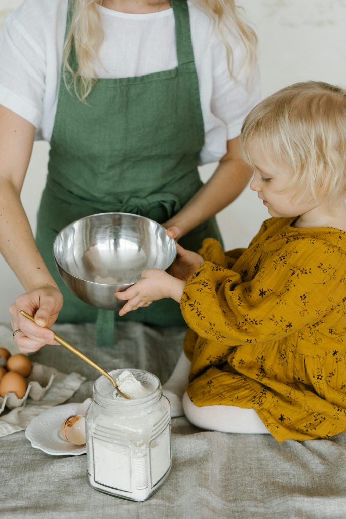 pexels-photo-6957990-6957990 High angle of little daughter sitting on table with stainless bowl helping mother in making dough